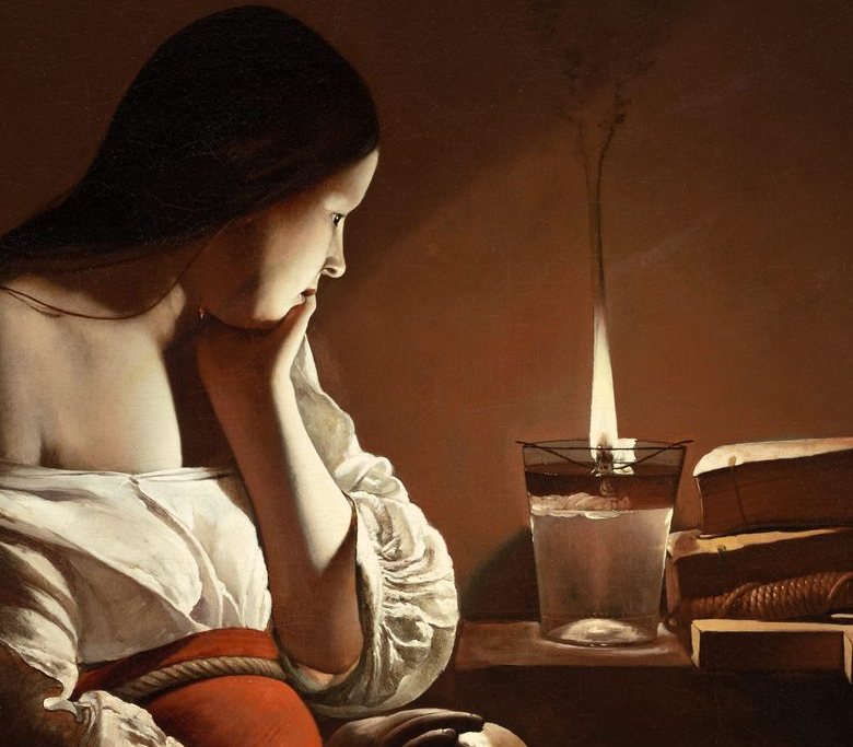 Detail of a flame burning on a floating wick in a glass of oil and water on a table, lighting the face, hand and white sleeve of a woman who gazes at it; everything farther away falls into brown darkness.